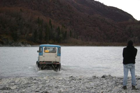 2003 Jeep TJ - A person watches a blue and white van drive through a shallow body of water near a rocky shore and wooded hillside.