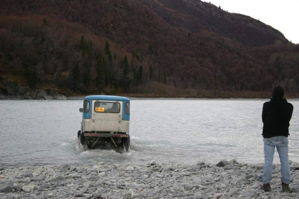 Blue and white van driving through shallow water with a person watching from shore