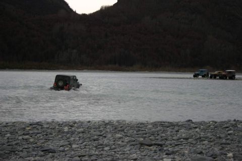 2003 Jeep TJ - Multiple off-road vehicles traverse a shallow riverbed near a wooded mountainside.