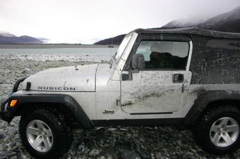 2003 Jeep TJ - A muddy Jeep Wrangler is parked on a rocky shore, with a glacial landscape visible in the distance under an overcast sky.