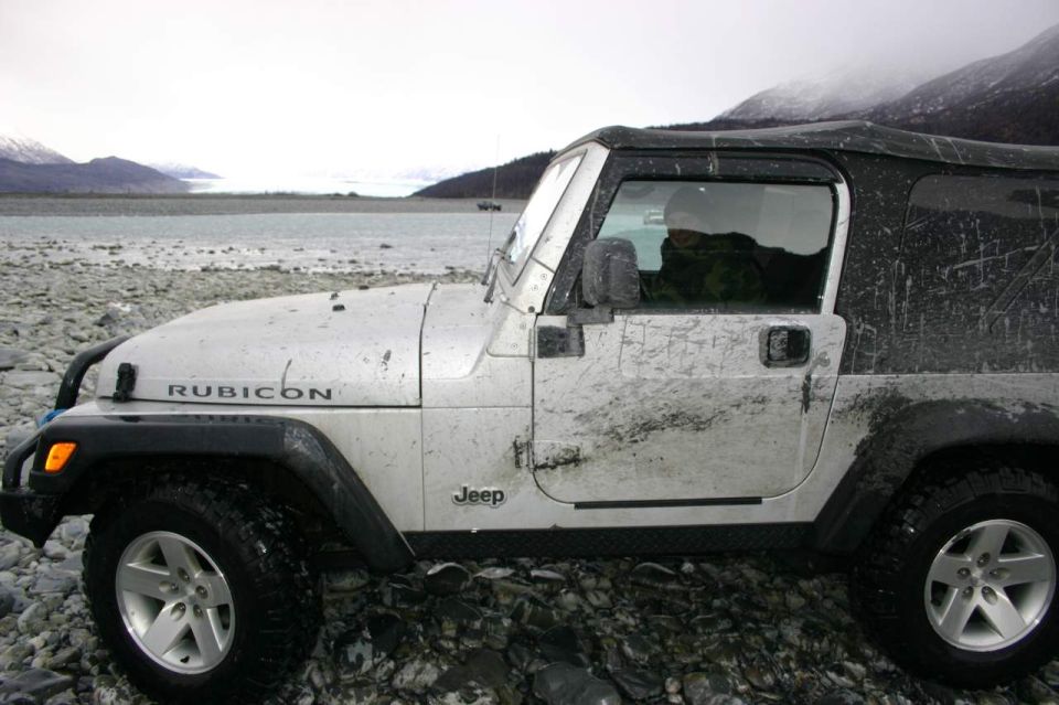 Side view of a silver Jeep Wrangler Rubicon covered in mud, parked on a rocky shore with mountains and a glacier in the background