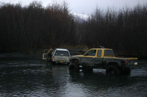 2003 Jeep TJ - Two trucks navigate shallow water surrounded by dense trees, with snow-capped mountains visible in the distance.