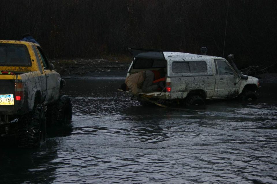 Yellow and white lifted mini-trucks axle-deep in a river crossing at dusk, person at the open tailgate