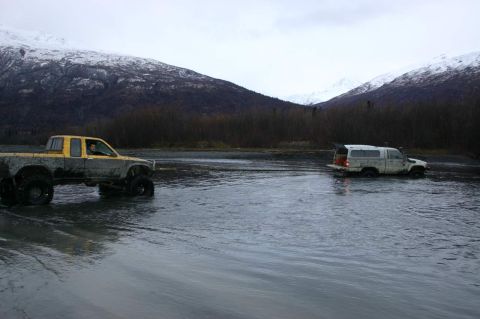 2003 Jeep TJ - Two trucks navigate a shallow body of water with snow-capped mountains in the distance under an overcast sky.