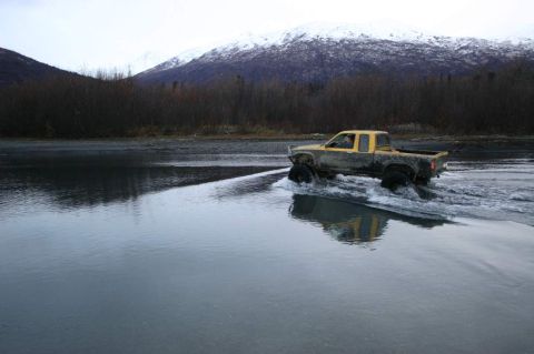 2003 Jeep TJ - A yellow pickup truck navigates a shallow river with snow-capped mountains in the background.