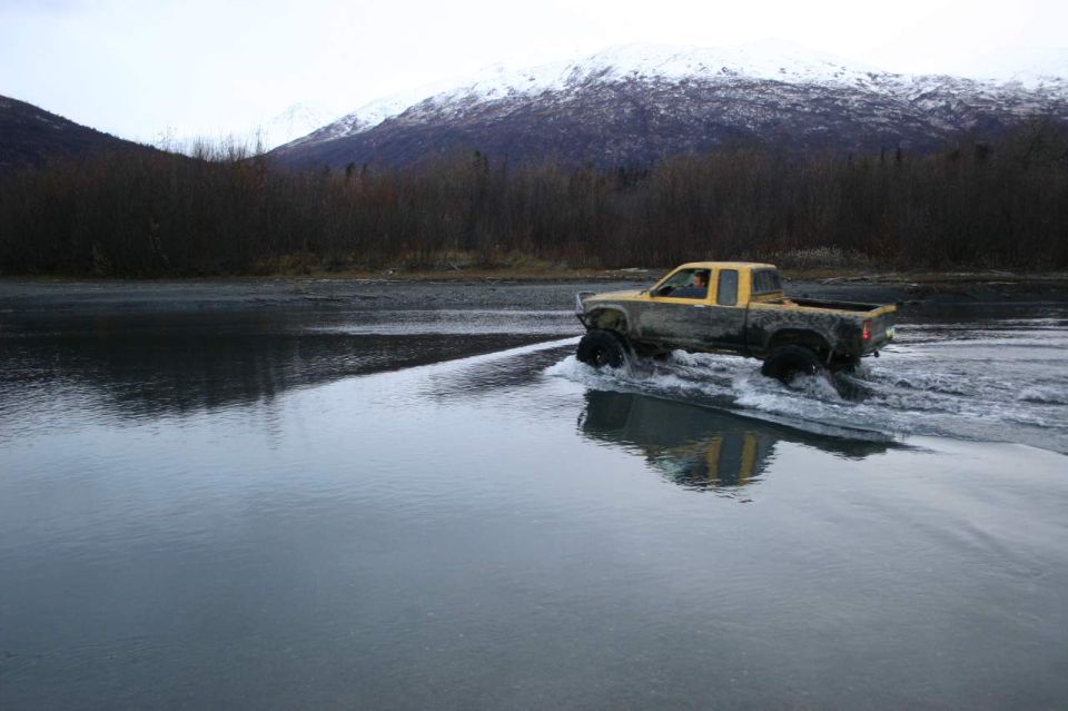 Yellow pickup truck with large tires driving through shallow water, mud splattered on body