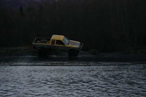2003 Jeep TJ - A yellow pickup truck with oversized tires navigates a muddy terrain near a body of water.