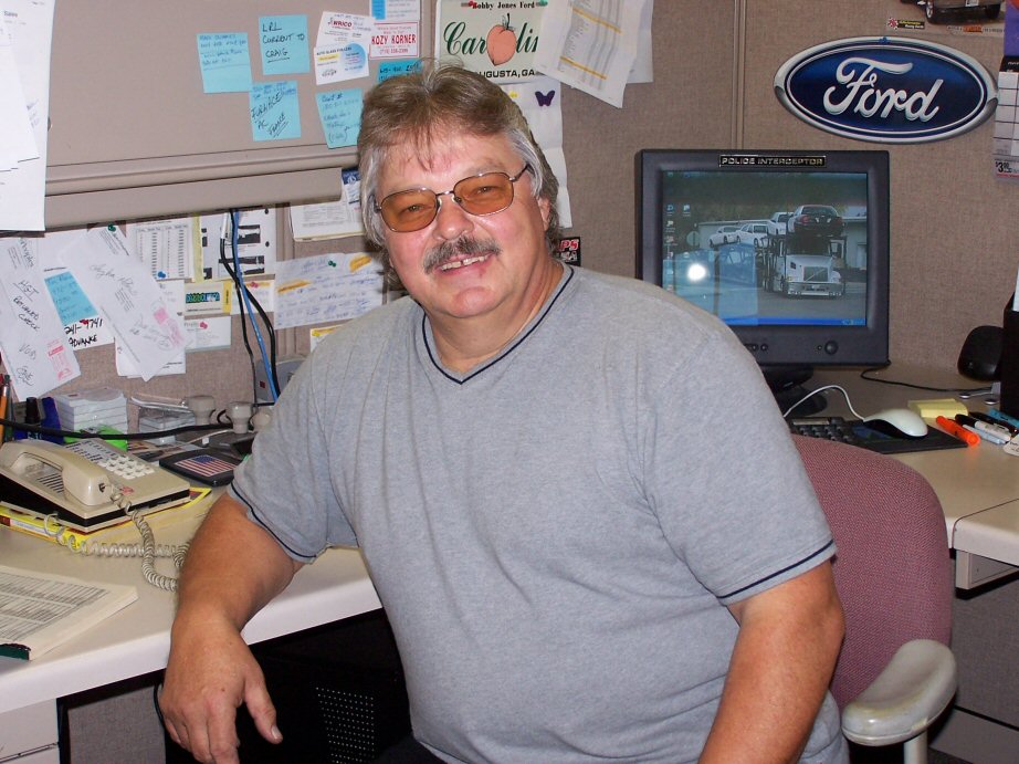 Mustached man in gray shirt seated at office cubicle desk under blue Ford oval sign