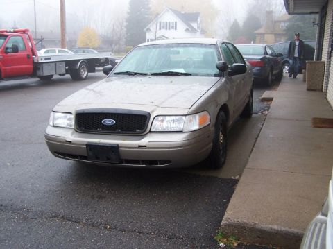 2001 Ford Crown Victoria - A tan 2001 Ford Crown Victoria is parked on a wet street next to a sidewalk, with a tow truck and other vehicles in the background.
