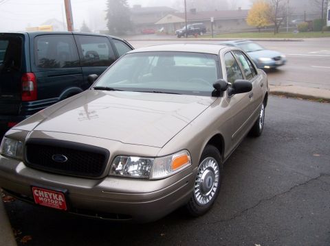 2001 Ford Crown Victoria - A tan 2001 Ford Crown Victoria is parked on a street next to a dark minivan, with a blurry car in the background.