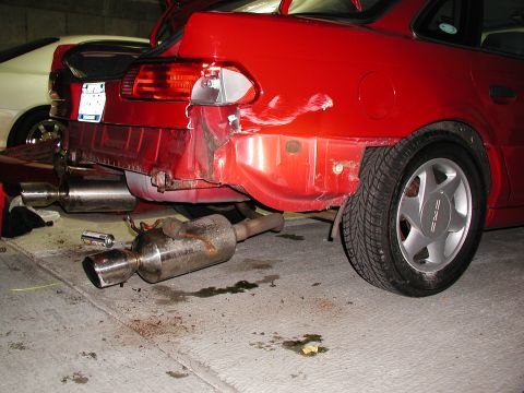 1992 Ford Taurus - A red car with a severely damaged rear bumper and a detached exhaust pipe lies on the ground. Another vehicle is partially visible in the b…