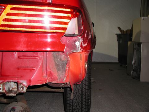 1992 Ford Taurus - Crumpled and torn red rear quarter panel showing crash damage beside the taillight and tire, photographed in a garage.
