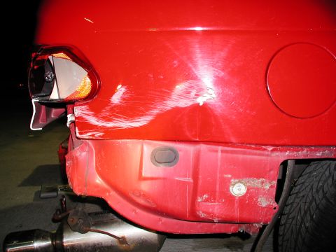 1992 Ford Taurus - The rear bumper of a red 1992 Ford Taurus shows extensive damage, including scratches, dents, and rust, with a broken taillight.