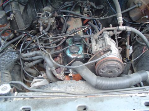 1986 Ford Bronco - A cluttered engine bay with various hoses and wires, a carburetor and AC compressor, with visible text at the bottom.