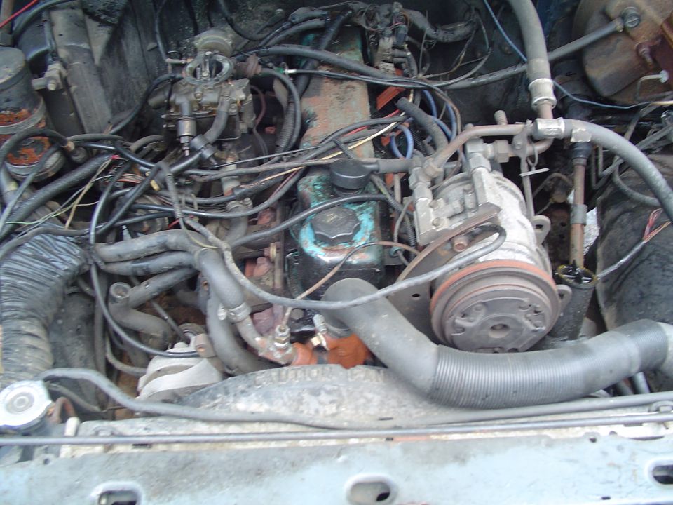 Engine bay of an older vehicle showing carburetor, AC compressor, hoses and dirty wiring