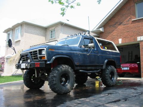 1986 Ford Bronco - This lifted 1986 Ford Bronco features aggressive off-road tires and auxiliary lighting, parked in a driveway next to a red sports car.