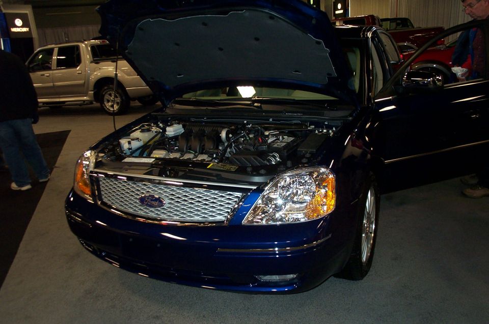Blue sedan's engine compartment and front grille are visible. A silver pickup truck is in the background