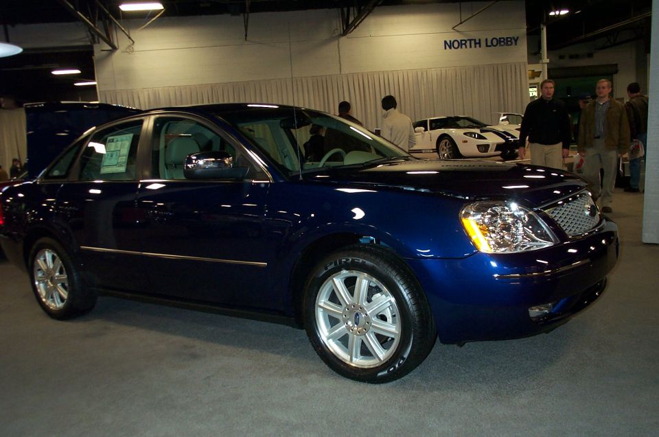 Dark blue sedan parked indoors, with a white sports car in the background. The sedan has chrome trim and multi-spoke wheels