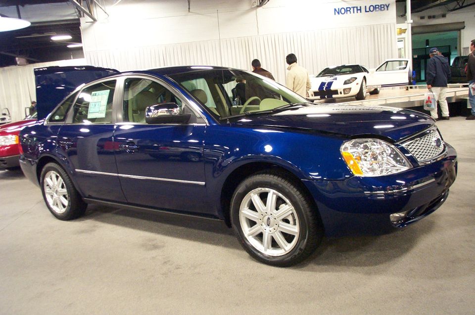 Blue sedan with chrome trim and multi-spoke wheels. A white Ford GT is visible in the background