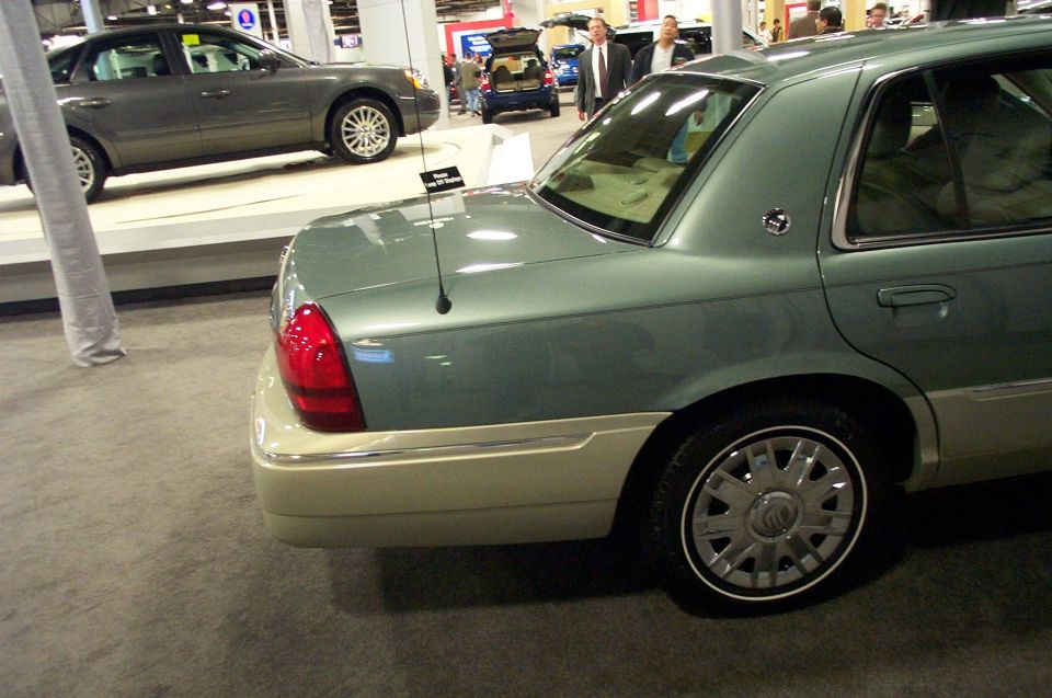 Rear quarter view of a light green Mercury Grand Marquis with a two-tone lower body. Another gray sedan is visible on a platform