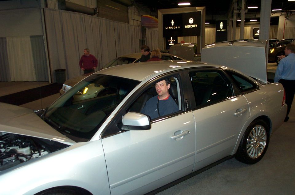 Silver sedan with hood open, driver inside, at an auto show