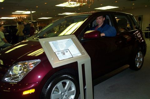 2001 Ford Crown Victoria - A man sits in a maroon hatchback parked indoors, with a display stand in front of the vehicle.