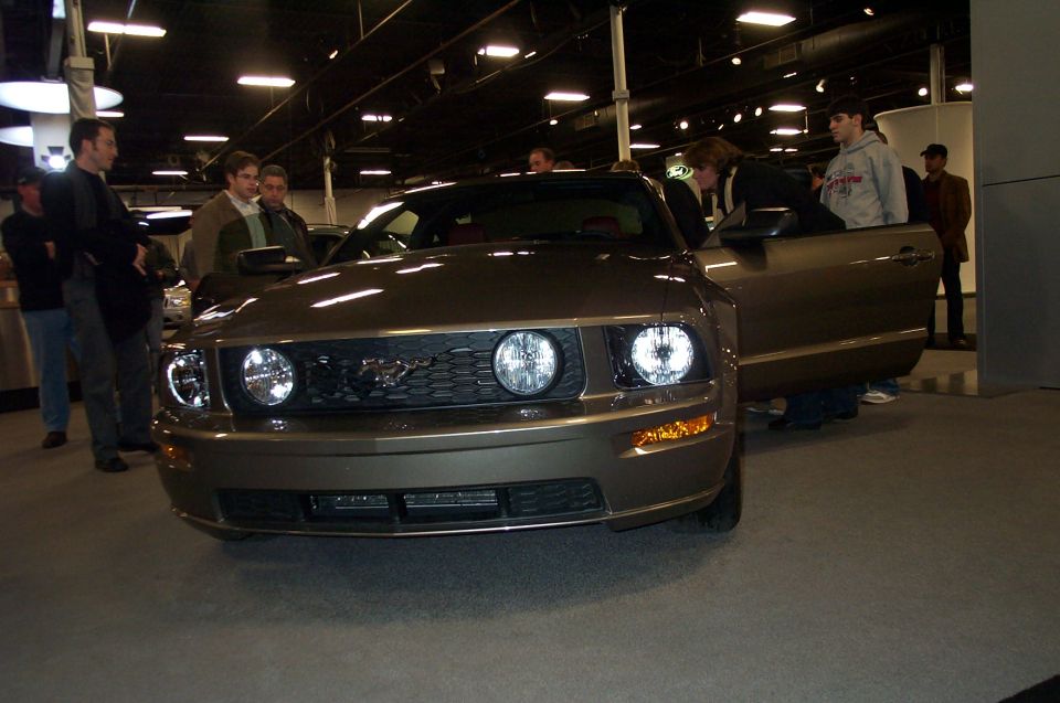 Front view of a brown Ford Mustang with its driver's door open. People are looking at the car