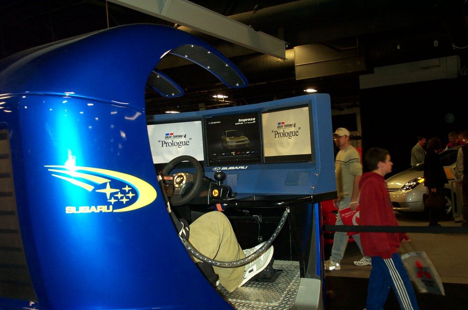 Blue Subaru racing simulator with screens showing Gran Turismo 4 Prologue