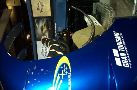 2001 Ford Crown Victoria - A person sits inside a blue Subaru Gran Turismo 4 driving simulator, with screens displaying game information.