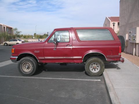 1991 Ford Bronco - This red Ford Bronco is parked in a lot with buildings and a light pole in the background.