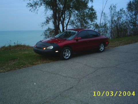 1997 Ford Taurus - A red sedan with a car bra is parked on the side of a road overlooking a body of water, with a timestamp indicating it was photographed on…
