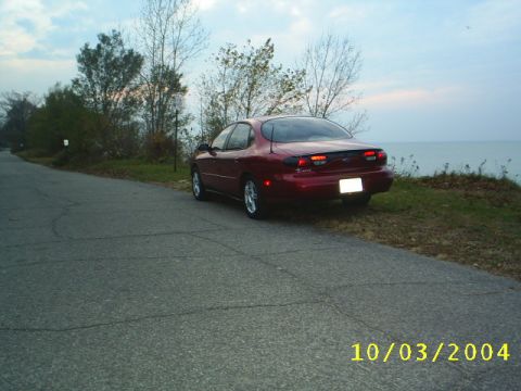 1997 Ford Taurus - A red sedan is parked on the shoulder of a road overlooking a large body of water under a cloudy sky.