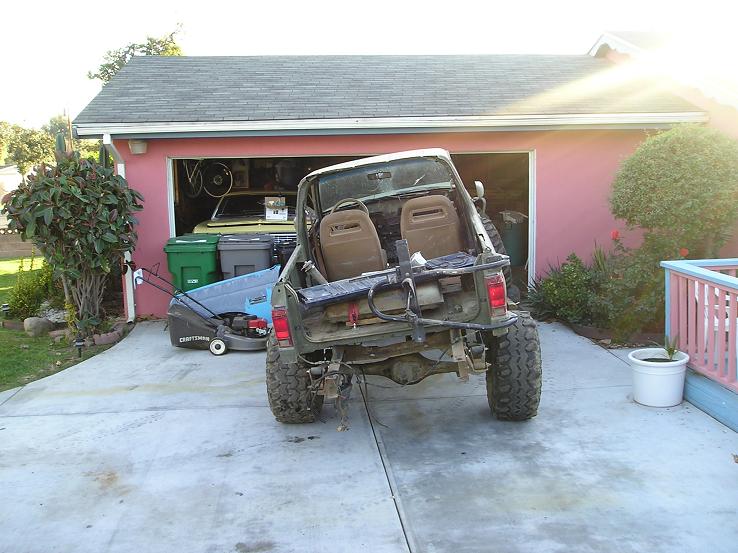 Off-road SUV with lifted suspension and large tires parked in front of a garage. It has a roll bar and two bucket seats visible
