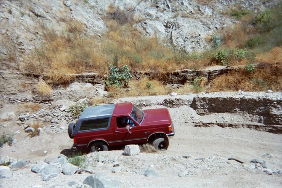 Red 1989 Ford Bronco off-roading on a dirt path with rocky terrain