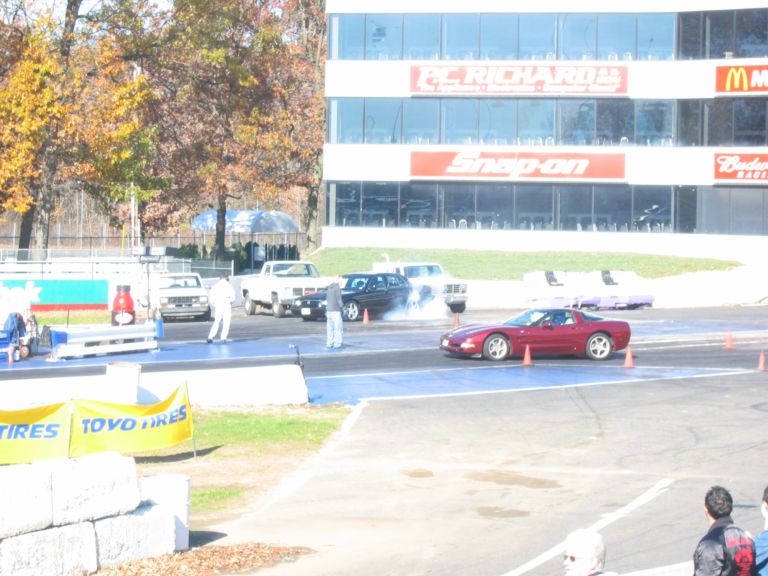 Dark sedan smoking tires at drag strip starting line; burgundy Corvette in foreground, sponsor-covered tower behind.