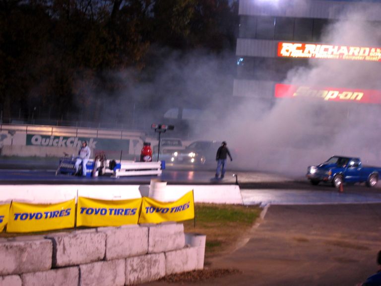 Heavy tire smoke at a drag strip starting line at dusk, with a blue pickup and Toyo Tires banners visible.