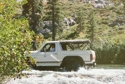 1994 Ford Bronco - A white Ford Bronco navigates a rocky stream surrounded by lush green foliage and distant hills.