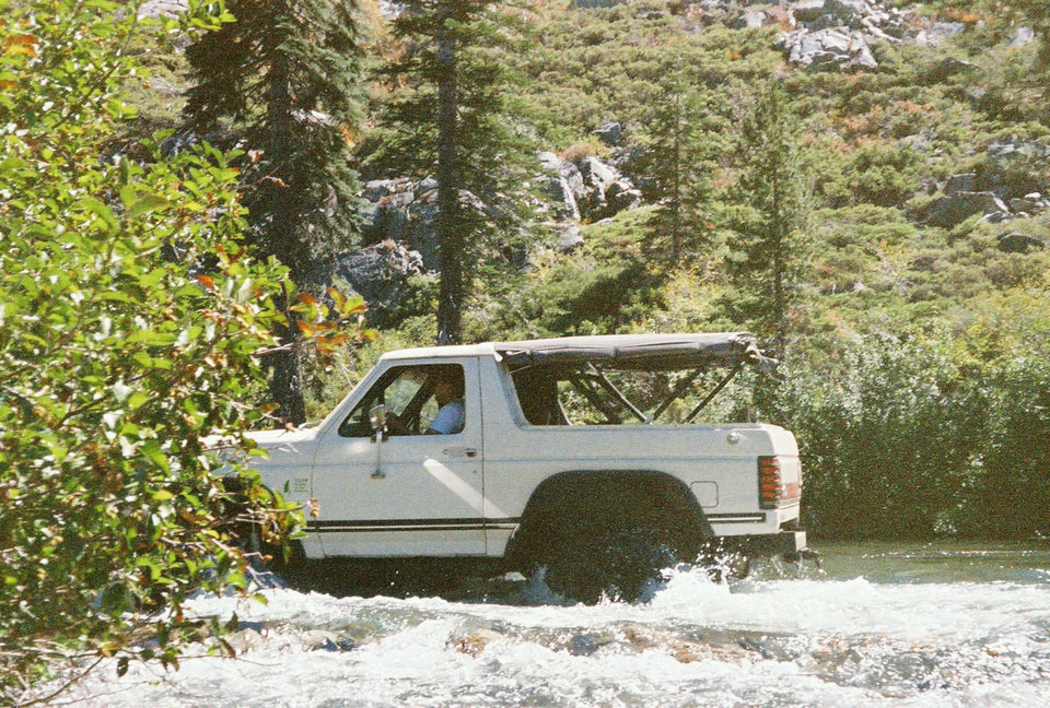 White two-door 4x4 SUV with top removed fording a rocky river