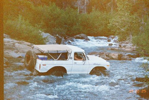 1994 Ford Bronco - A white 1978 Ford Bronco navigates a shallow, rocky river with trees and brush lining the banks.
