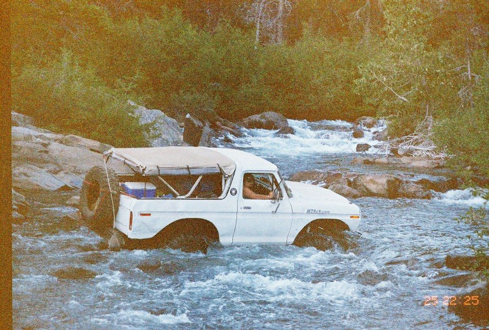 White doorless Ford Bronco with tan soft top fording a whitewater river