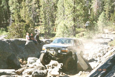 1994 Ford Bronco - Ford Bronco tackling a challenging off-road course, with spectators watching from a rocky outcrop.
