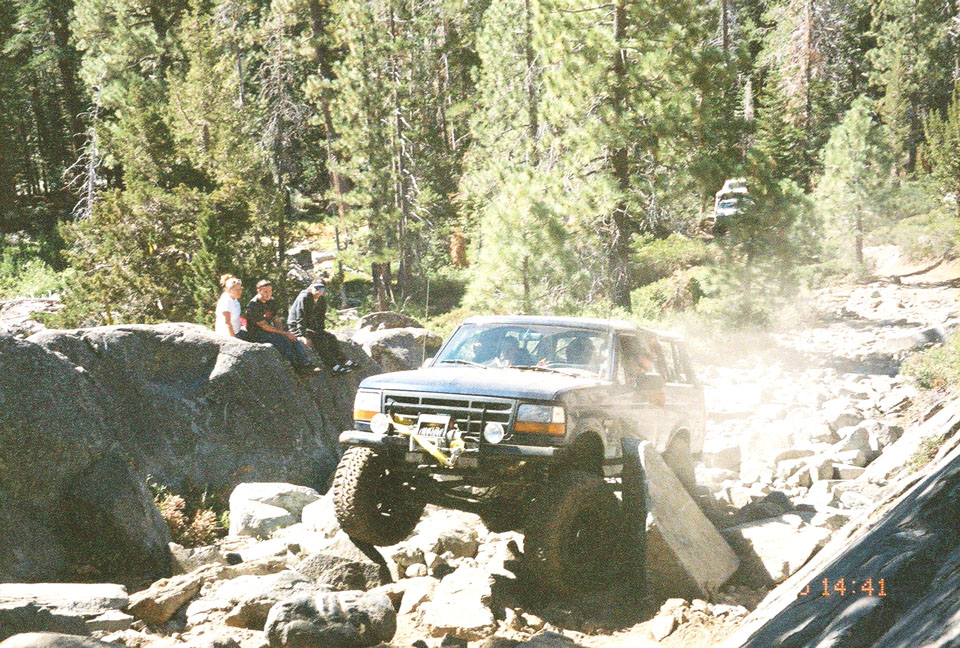Modified Ford Bronco navigating a difficult rocky trail with another vehicle visible in the background