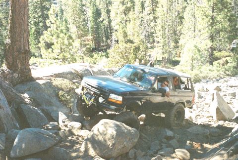 1994 Ford Bronco - A driver maneuvers a black Ford Bronco over a challenging rocky terrain in a wooded area.