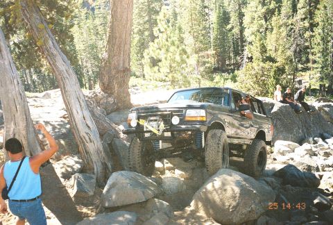 1994 Ford Bronco - A black Ford Bronco is driven over large rocks on a trail, with people watching from a boulder.