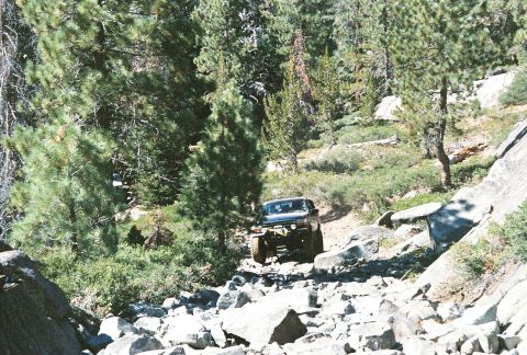 1994 Ford Bronco - A dark-colored SUV is seen off-roading on a challenging, rocky trail surrounded by trees and large boulders.