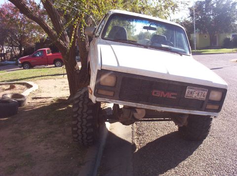 1988 Ford Bronco - White GMC pickup truck with oversized tires is parked on a curb, with a red pickup truck visible in the background.