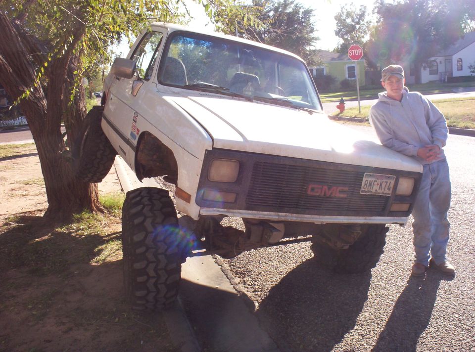guy leaning on front of white GMC pickup truck with front tire up in a tree