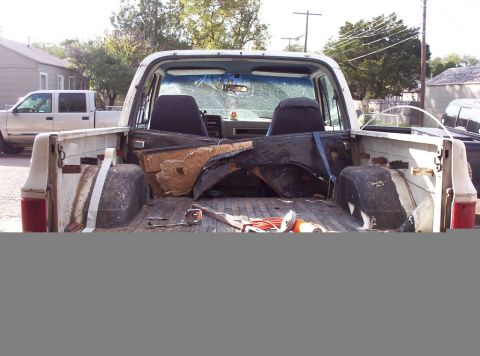 1988 Ford Bronco - The interior of this white pickup truck bed has been removed, revealing the wooden floor and various dismantled parts. A white SUV is visib…