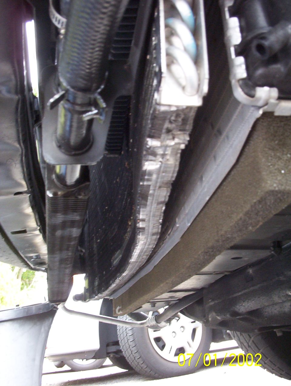 Under-hood view down onto radiator and condenser stack with hoses, front tire and wheel cover below, dated 07/01/2002