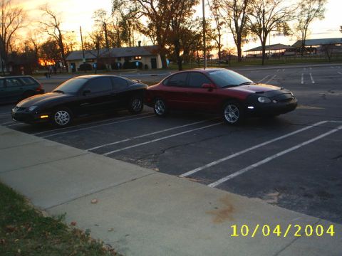 1997 Ford Taurus - Two cars, a red 1997 Ford Taurus and a black car, are parked in a lot at sunset.
